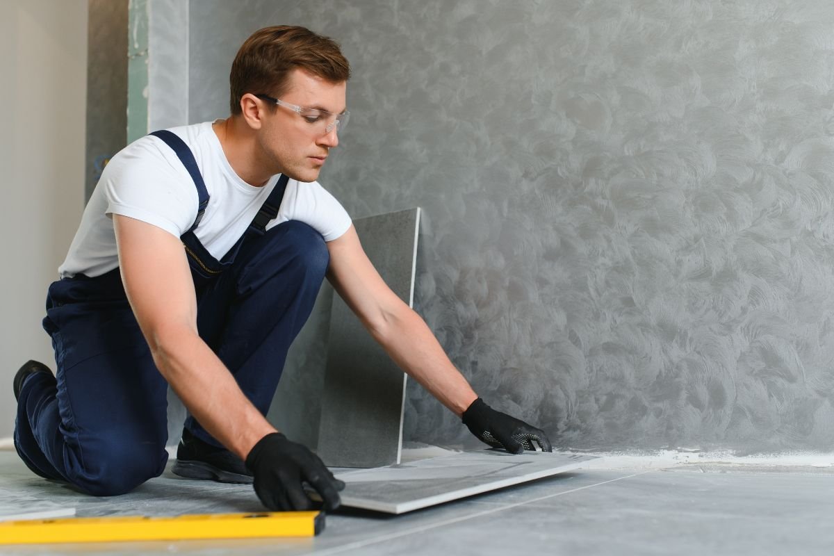 A person kneels on the floor, wearing safety glasses and gloves, positioning a tile against a gray textured wall. A yellow level tool lies nearby.