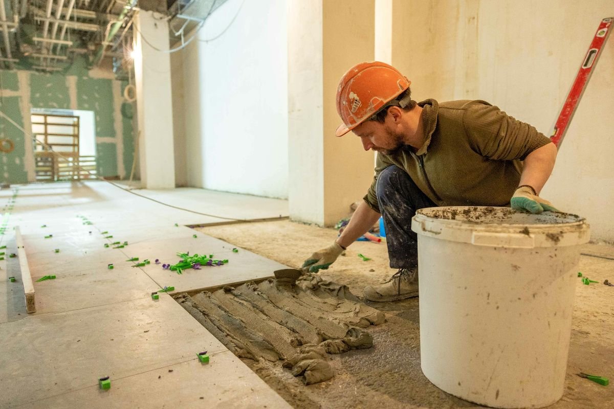 A construction worker wearing a hard hat is kneeling on the ground, applying mortar with a trowel to lay tiles in an unfinished building interior. A bucket and scattered tools are nearby.